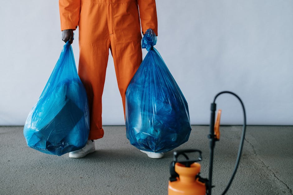 A person dressed in an orange protective jumpsuit and white footwear is standing on a smooth concrete surface, holding two large blue rubbish bags filled with waste, one in each hand. The bags appear 