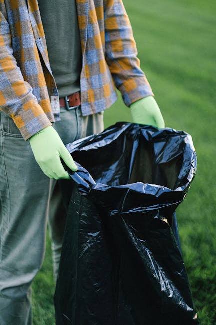 A person wearing a green long-sleeve shirt, grey pants, and yellow-and-blue checkered jacket is holding open a large black rubbish bag lined with a silver interior, positioned outdoors on green grass.