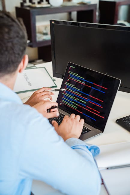 A person using a laptop placed on a dark surface, with their hands positioned over the keyboard. The screen displays lines of code with syntax highlighting in various colors such as green, blue, and w
